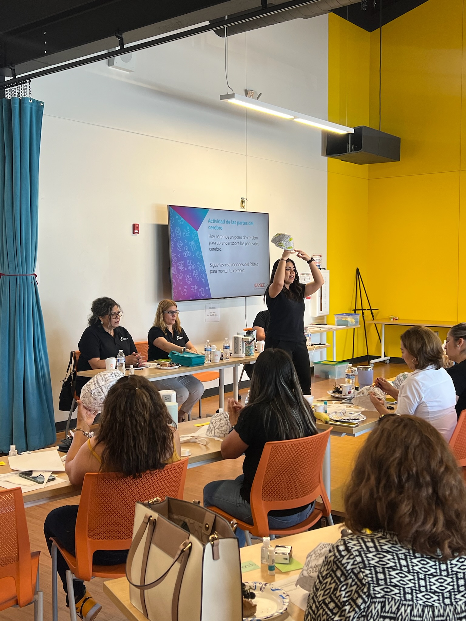 Jessica teaching a class in a yellow room, holding up a hat